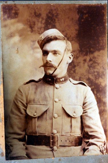 This image is a portrait of a soldier, taken from the waist up.  He is dressed in army uniform.  His jacket is buttoned up to the neck and a wide leather belt is around his waist.  His small pillbox hat is sitting on the right-hand side of his head and has a leather chin strap.  He has a large moustache, a serious look on his face and is looking away from the camera.  