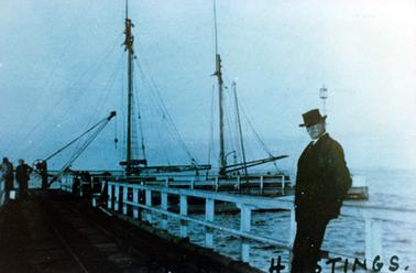 This image shows a two-masted boat at the end of a pier.  A man, dressed in a dark coloured suit, white shirt, tie and hat, is leaning against the pier railing.  He is looking at the camera.  A small crane is at the end of the pier.  Two men are standing at the end of the pier on the left-hand side.  Rail tracks are running down the centre of the pier.  Written in black ink at the bottom right-hand side of the image is “Hastings”.  
