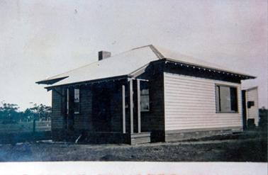 This image shows a small weatherboard house with a sloping roof at the front.  A small verandah is across the front of the house with a window on either side of the front door.  The side wall has one window at the right-hand end.  Part of a small building can be seen at the back of the house.  Grass is in the foreground and a paddock with tall trees can be seen in the background on the left-hand side of the house.  