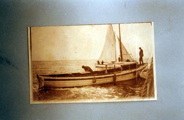 This image shows two boats in the water.  The wooden boat in the foreground is long and narrow and has a small cabin.  Two men are on board.  The boat in the background, has two sails and two men can be seen on board.  Part of a rope can be seen in the near right-hand corner.  