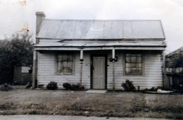 This image shows a small timber cottage with a sloping roof and a four posted verandah across the front.  A large window sits on either side of the front door.  A brick chimney is on the side wall on the left-hand side of the building.  A fence can be seen at the back on either side of the house in front of bushy shrubs.  Part of a roof of the house next door can be seen on the far right-hand side.  Small shrubs are planted in front of the verandah and a grassed lawn and a gravel road are in the foreground.