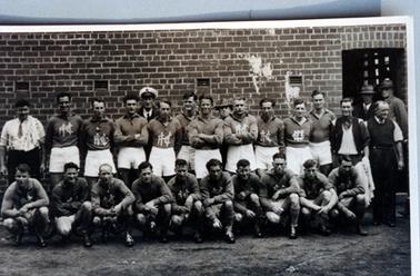 This image shows a football team, posing for a photo.  There are ten players standing and ten players crouching on the ground at the front.  A man in a white shirt and tie with a towel over his shoulder, is standing on the far left-hand side and a man in a dark coloured shirt, tie and white sleeveless jumper, holding a towel, is standing on the right-hand side.  Three other men are standing at the right and two men, one with a sailor’s hat are standing at the back.  A brick wall forms the background with an open doorway on the far right-hand side.  