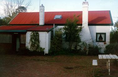 This image shows a white weatherboard cottage with a red corrugated iron roof.  Two white chimneys are at the front of the house.  A small porch with lattice is across the front is on the left-hand side of the cottage.  Shrubs and creepers are across the front of the house and a picnic table is on the lawn.