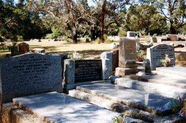 This image shows a cemetery with four graves with large headstones in the foreground.  Many headstones can be seen in the background and tall trees are in the far background.  