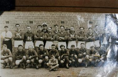 This image shows a football team, posing for a photo.  There are ten players standing and ten players crouching on the ground at the front.  A man in a white shirt and tie with a towel over his shoulder, is standing on the far left-hand side and a man in a dark coloured shirt, tie and white sleeveless jumper, holding a towel, is standing on the right-hand side.  Two men, one with a sailor’s hat are standing at the back.  A young boy is sitting cross-legged on the ground at the front of the group.  A brick wall forms the background with an open doorway on the far right-hand side.  