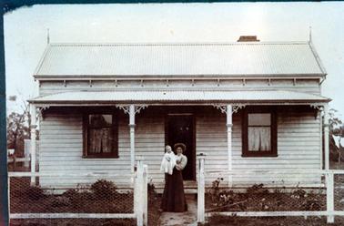 This image shows a woman holding a baby, standing in front of a weather-board cottage, posing for a photo.  They are both beautifully dressed.  The woman is wearing a white high necked, long sleeved blouse and a long dark coloured skirt.  She has a mop of dark coloured hair.  The baby is dressed in a beautiful long white dress.  The cottage has a four post verandah across the front.  A single window with lace curtains, is on either side of the front door.  A timber and wire netting fence is across the front and small shrubs are on the front lawn.  Part of a clothes line is on the far right-hand side.  