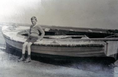 This image shows a boy sitting on the edge of a wooden fishing boat, posing for a photo.  He is dressed in dark coloured shorts and a pullover, shoes and socks.  His feet are resting on the ground.  His fair hair is parted on the left side.  
