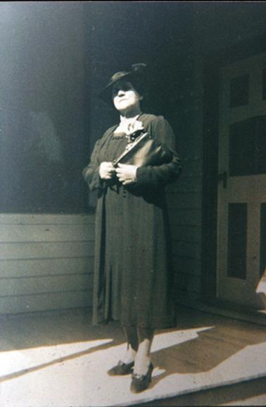 This image shows a well dressed matronly woman standing on a timber deck in front of a panelled door.  She is wearing a dark coloured dress with matching full length coat.  She is wearing a hat, court shoes and is holding a handbag under her left arm.