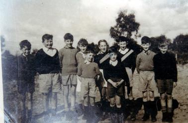 This image shows a group of ten boys standing in a paddock, all smiling for the camera.  All the boys are dressed in shorts, jumpers and shoes and socks.  Trees are in the background.  