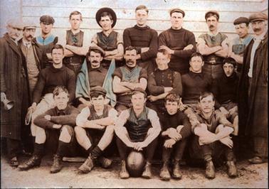 This image shows a group of twenty-one men, standing and sitting in three rows, posing for a photo.  There are ten men standing at the back, six men sitting in the middle row and five men sitting on the ground.  Ten men are wearing caps and one man in the back row is wearing a hat on the back of his head. The two older men standing on either side are wearing long coats.  The man at the centre of the front row has a football between his feet.  A weatherboard building is behind the group.