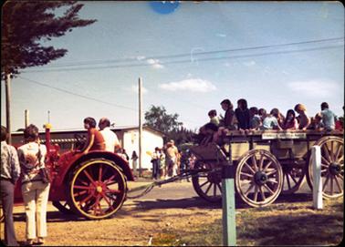 This image shows a red tractor pulling a wooden four-wheeled wagon filled with children.  There are approximately a dozen children in the wagon and a man and child are on the tractor.  People are standing on either side of the road watching.  Part of a building can be seen in the background on the left-hand side and the branches of a pine tree can be seen in the top left-hand corner.  Two guide posts are in the foreground on the right-hand side of the image.  