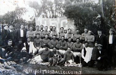 This image shows a large group of thirty-six footballers and other men and children, posing for a photo.  There are eighteen men in the back row, six players sitting on chairs in the middle row and twelve men and boys sitting and kneeling in the front row.  A small boy is sitting on the lap of a player in the centre of the photo, holding a football with “H.F.C. 1913” printed on it.  A large banner with “H.F.C.” printed in large letters is behind the group.  Tall trees are in the background.  A pile of pipes are in the foreground on the far left-hand side of the image.  Printed across the bottom of the photo in white ink is: “HASTINGS FOOTBALL CLUB 1913”.