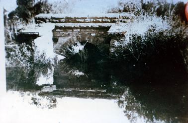 This image shows an old stone bridge with an arch underneath, across a creek.  The reflection of the bridge can be seen in the water.  Bushes are on either side of the creek and tall trees can be seen in the background.  