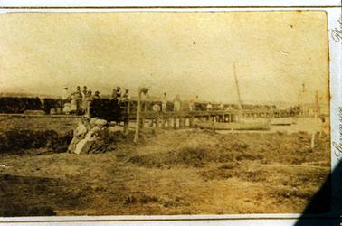 This image shows a long wooden pier with a group of men and children standing at the left and several men standing along the jetty.  A group of four women wearing long dresses and large hats, are sitting on the shore in front of the jetty.  The woman on the left is holding a baby.  Fishing boats can be seen on the right-hand side of the jetty and the foreshore is in the foreground.  Fishing nets are hanging along the rail behind the people.