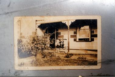 This image shows three small children standing on the verandah of a brick house.  A turned verandah post with lace wrought iron at the top, is in the middle of the image.  A front door is at the left and a large, single window is to the right of the door.  A garden and shrubs are on either side of a path.   