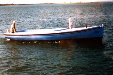 This image shows a couta boat on the water with a man sitting in the stern, looking at the camera.  Land can be seen the far background.  