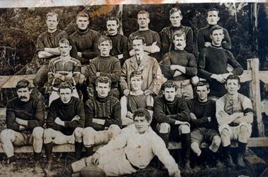 This image shows a football team posing for a photo.  There are six men sitting on the top railing of a fence, five men standing in the middle row and seven men sitting on the bottom rail of the fence.  One man is half lying on the grass at the front.  All the players have arms crossed except the man at the centre of the middle row who is dressed in a white shirt, tie and coat and has his hands resting on the shoulders of the man in front of him.  Trees and shrubs are in the background.