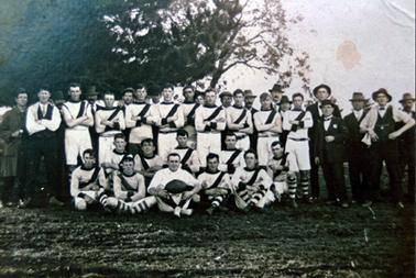 This image shows a group of thirty-five men standing and sitting, in four rows, posing for a photo.  There are eighteen players dressed in long white shorts, white shirts with a black band across the front and black and white striped long socks.  A man dressed all in white and holding a football is sitting at the centre front.  Several men, dressed in suits, hats and caps, are on either side and behind the players.  A large tree is behind the group.  