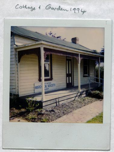 This image shows the front of a timber cottage.  Two windows are on either side of the panelled front door.  A small verandah is across the front held up by three posts.  A lacy wrought iron trim surrounds the verandah posts.  An empty garden bed and a brick pathway are in front of the cottage.  A sign is attached to the front wall which reads: “Shire of Hastings Historical Society Project”.