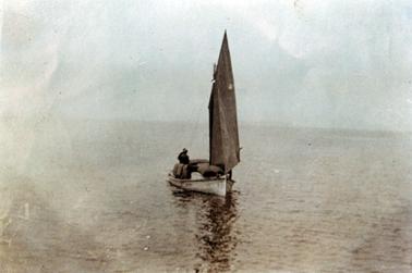 This image shows a small wooden fishing boat with a tall sail.  The boat is in the centre of the image, surrounded by water.  The top half of two men can be seen with their backs to the camera.  