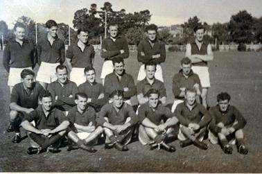 This image shows a football team, standing and sitting on an oval, posing for a photo.  There are six men standing in the back row, six men squatting in the middle row and six men sitting, crossed legged in the front row.  The players are all wearing white shorts and dark coloured long sleeved shirts except for the player on the far right in the back row, who is wearing a sleeveless shirt.  Tall trees and houses can be seen in the background, behind a fence.