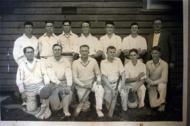 This image shows a group of twelve cricket players standing and kneeling in two rows, plus a man dressed in a white shirt, tie and suit who is standing at the far right-right hand side in the back row.  There are six players standing in the back row and six players kneeling in the front row.  The two players in the middle of the front row are holding cricket bats.  Another player is kneeling on a cricket pad.  A timber building is behind the group.  