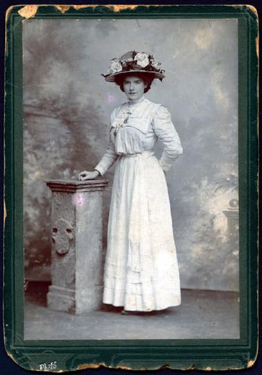 This image shows a full length portrait of a woman taken in an photographer’s studio.  She is standing with her right hand resting on a pillar and her left hand behind her back.  She is wearing a white full length dress with long sleeves and a large square collar.  A locket is around her neck.  Thick dark coloured hair can be seen underneath a large hat, which is decorated with large flowers.  The word ‘Photo’ is printed in silver at the bottom of the mount.
