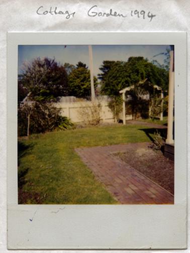 This image shows a small front and side lawn and garden.  One verandah post of a building can be seen on the far right-hand side of the image.  An empty garden bed, a brick path and green grass are in the foreground.  A white picket fence with an arbour runs across the front of the property.  A wooden fence with tall trees behind it can be seen in the background.  Part of an electricity post can be seen behind the picket fence.  