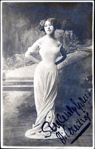 This image shows a young woman posing for a photo in a photographer’s studio.  She is wearing an off the shoulder white dress with a tight fitting bodice and a long skirt which is twisted and spread out on the floor at her feet.  Her thick wavy hair is parted in the centre and she is looking away from the camera.  Her hands are behind her back.  A scene is on the studio wall behind her.  Written across the bottom of the photo in large back writing are the words “Sincerely Yours Vi Coudrey”.