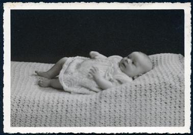 This image shows a baby lying on a baby blanket on a bed.  The baby has bare feet and is wearing a white knitted dress with ribbon at the front. The photo has a serrated edge around the white border.   