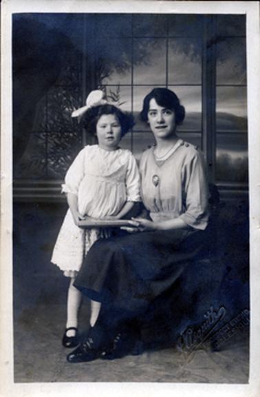 This is a studio portrait of a woman and small girl. The woman is sitting on a chair holding one side of a book in her left hand.  She has dark coloured hair parted on the right, a light coloured blouse with three-quarter sleeves and a dark coloured skirt.  She is wearing black stockings and black lace-up shoes.  She has a large brooch in the centre of her blouse and a small chain around her neck.  The little girl is standing beside her and holding the other side of the book in her right hand.  She is wearing a white knee-length dress, long white socks and black shoes.  She has a large white bow in her dark coloured hair.  The studio backdrop has a scene with windows in the foreground.  The photographers details are embossed at the bottom right corner of the image.  