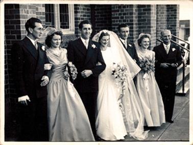 This image shows a wedding party standing outside a brick building posing for a photo.  The bride and groom are in the centre and a bridesmaid and bestman/groomsman are on either side.  The brides father on on the far right-hand side of the group.  