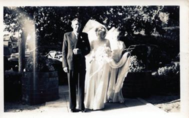 This image shows a bride and her father arriving at the church before the wedding ceremony.  The bridesmaid is behind the bride, holding the bride’s train.  Brick garden ‘pots’ are on either side of the pathway.  Wedding cars can be seen behind the group as well as trees and shrubs.  