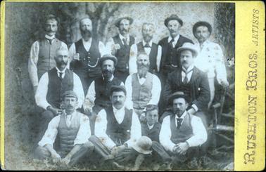 This aged and faded image shows a group of thirteen men and one boy, sitting and standing in three rows, posing for a photo.  There are six men standing in the back row, four men sitting on chairs in the middle row and three men and one boy sitting cross-legged on the ground.  Most of the men are wearing long-sleeved white shirts and waistcoats.  Two of the men and the boy are also wearing suit coats.  Most are wearing either a hat or a cap.  The man at the back on the far right-hand side has his hand resting on a chair.  “Rushton Bros Artists” is printed across the far right-hand side of the photograph.  