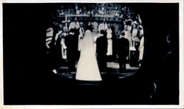 This small oval image shows a wedding service taking place inside a church.  The image is surrounded on three sides by a black background. The photo is not in focus.  The bride and groom and two men have their backs to the camera.  Other people, including the minister, can be seen standing.  A cross is standing on the left-hand side of the image.  