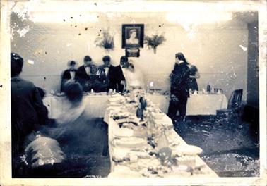 This image shows two long tables, arranged in a T-shape, set for a wedding reception.  The bride and groom are standing at the centre of the top table and appear to be cutting the wedding cake.  Three men are standing behind the table on the left-hand side and and man and a woman are standing on the right-hand side.  A framed photo of the queen is on the wall above the bridal couple. The image is very blurred and the photo has white patches all over it.  