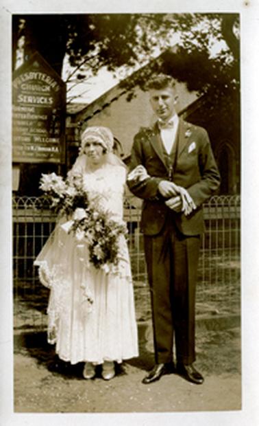 This image shows a very short bride and a very tall groom standing in front of a church, posing for a photo.  The bride is holding a large bouquet of flowers and has her left arm around the groom’s elbow.  The groom is wearing a three piece suit and is holding a pair of gloves in his right hand.  They are standing in front of a wire fence and a notice board, large trees and part of the church is in the background.  The notice board has ‘Presbyterian Church Services’ printed at the top and the service times and church particulars are printed beneath.  