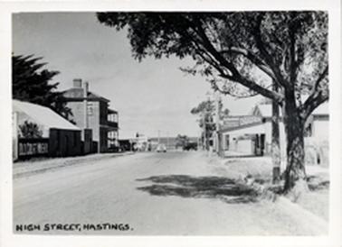 Black and white photograph showing a tree in the foreground on right and buildings along this side of the road. The draper’s store is one of them. There is a car in distance along street. On the left are a series of buildings including a double storied hotel; a large pine tree is situated beside the hotel. In print at bottom left-hand side are words “High Street Hastings”