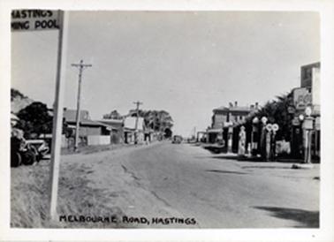 This image shows a street scene with businesses on either side.  A line of petrol bowsers is on the right-hand side.  A motor car is in the middle of the street coming towards the camera.  Electricity poles line the left-hand side of the street.  A sign post with “Hastings Swimming Pool” is in the foreground.  Printed across the bottom of the image is: “Melbourne Road, Hastings”.