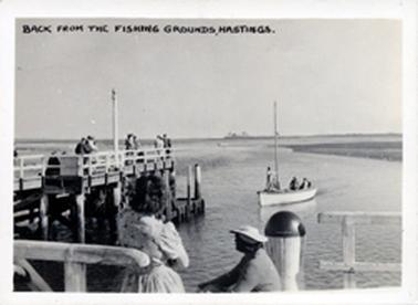 This image depicts a wooden boat with a tall mast returning to the jetty. Three people are on board.  There are a number of people standing on the pier.  The backs of a man and woman are in the foreground.  Land can be seen in the far distance.  Printed across the top of the photo is: “Back from the fishing grounds, Hastings”.  