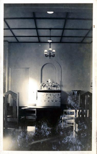 This image shows a church interior.  A cross sits on the altar.  The alter is covered with an embroidered cloth.  Tall-backed wooden chairs are in the foreground.  The ceiling has dark coloured timber squares and a pendant light hands from the ceiling at the centre of the image.  