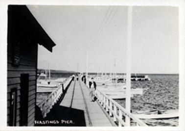 This image shows a jetty running out into the water.  Rail tracks run down the centre of the jetty and a white timber railing is on either side.  Part of a shed can be seen in the foreground on the left-hand side.  Fishing boats are on either side of the jetty.  People can be seen walking along the jetty.  Land can be seen in the far distance.  