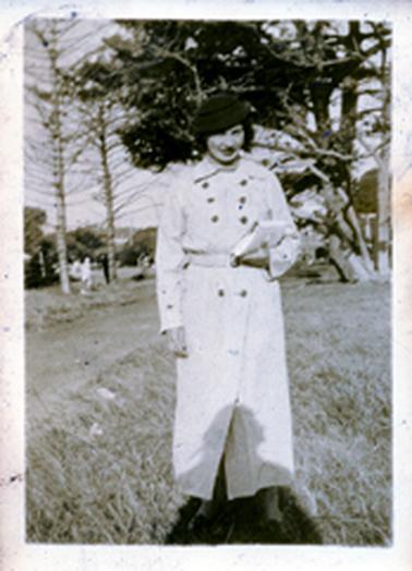 This image shows a woman standing on untidy grass, posing for a photo.  She is dressed in an ankle length striped, double-breasted coat.  She is wearing a small dark coloured hat and is holding a clutch bag in her left hand.  A pathway is on her right, leading down to a group of people.  Tall pine trees are in the background.