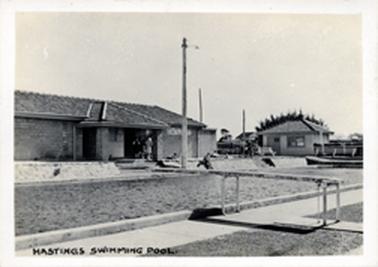 Black and white photograph of a swimming pool featuring the diving board in the foreground. Two buildings are depicted in the background.  People can be seen sitting and standing on the far side of the pool.  A tall light pole is at the centre of the photo.  Grass is in the foreground.  