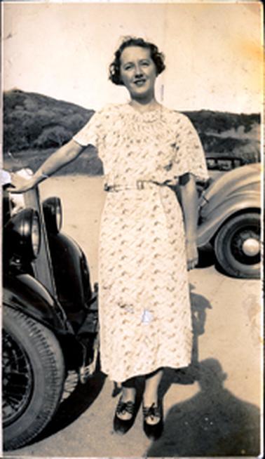 This image shows a young woman standing, posing for a photo. Her right hand is resting on the front of a motor car.  Only the two headlights and part of a wheel can be seen.  Part of another car can be seen to her left.  She is wearing a short sleeve print dress and black shoes tied with a bow on the top of her feet.  She has short dark coloured hair and has a smile on her face.  Trees and a hill can be seen in the background.  