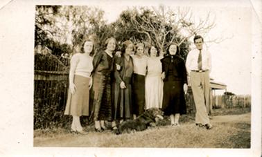 This image shows a group of six women and one man standing in a line, posing for a photo.  The woman are standing close together with arms linked or hands on shoulders.  The man at the right-hand end of the group is wearing a white shirt, tie and long trousers.  His hands are behind his back and his feet are crossed.  A large dog is lying at the feet of the women.  The group is standing in front of a fence and large trees are behind them.  Part of the verandah of a house can be seen on the far right-hand side.  