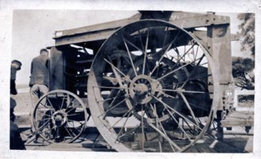 This image is a side-on view of an early model tractor showing a very large back wheel and a smaller front one.  The wheels are steel and have spokes.  Part of the engine can be seen behind the wheels and the backs of two men can be seen at the front of the tractor.  Tree branches can be seen on the far right-hand side.   