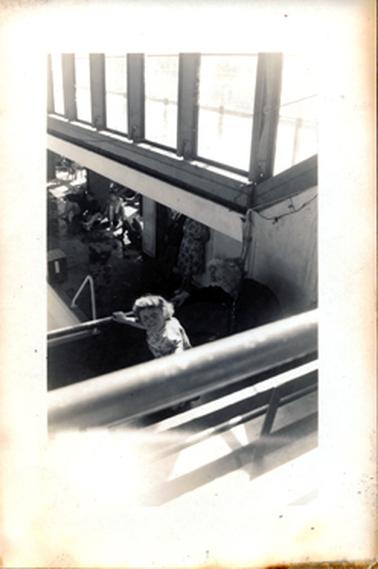 This image shows a young girl and an elderly woman holding onto a rail and looking up at the camera.  People are sitting and standing on the floor below them and a wall of windows are above.  A rail is in the foreground.  