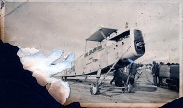 This badly damaged photo shows a small wingless aeroplane standing on a roadway.  A long road with a fence on either side leads to buildings in the distance.  A man is standing on the right and the legs of another man can be seen underneath the plane.  The letters ‘VH-UMN’ are printed on the side. The left-hand side of the photo has been torn away.  