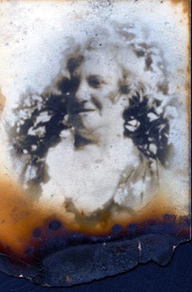 This badly damaged photo shows the head and shoulders of a woman.  She has a slight smile on her face and her head is turned to the left.  Her hair is fair.  Tree branches can be seen behind her.  
